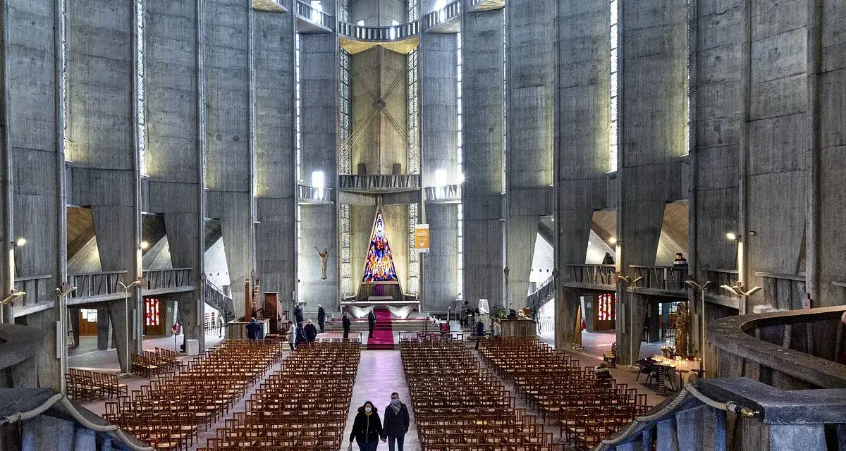 Intérieur de la cathédrale de Royan, avec ses murs en béton brut, ses rangées de chaises et son vitrail coloré au fond.