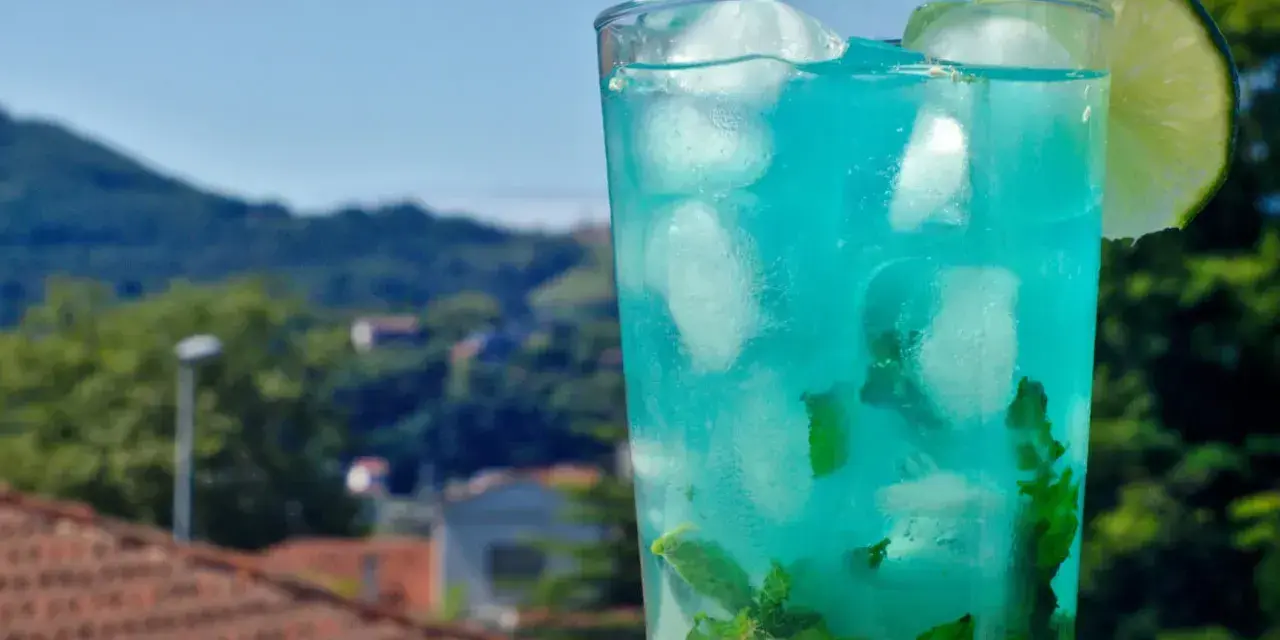 Orzeźwiający drink z blue curacao i sprite, z miętą i lodem, ozdobiony plasterkiem limonki, na tle zielonych wzgórz.