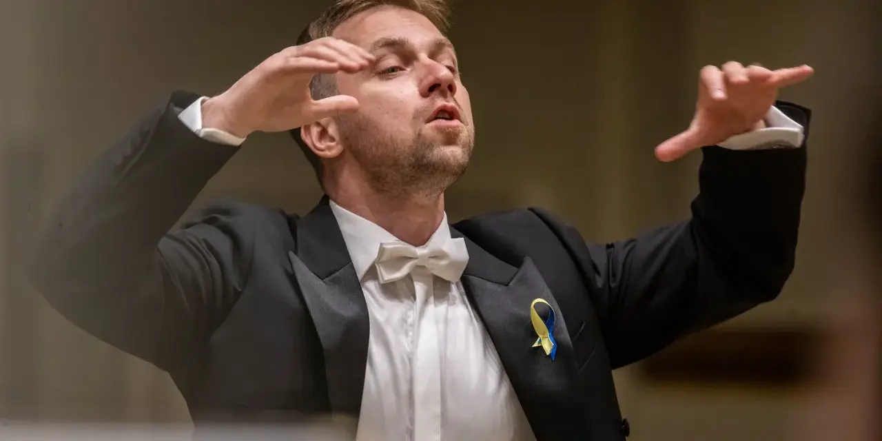 Dyrygent w garniturze z kokardką, prowadzący wykonanie Carmina Burana, z ukraińską wstążką na klapie.