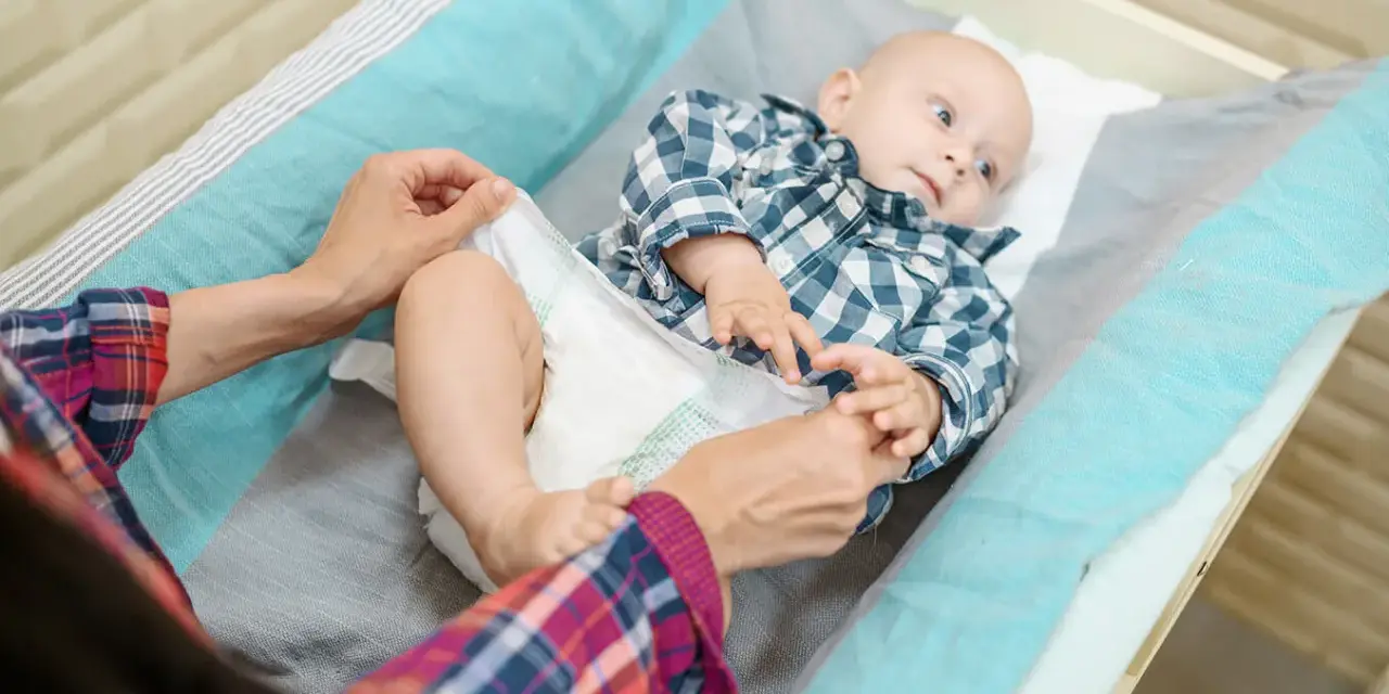 Mama sprawdza, kiedy zmienić rozmiar pieluchy maluszkowi. Dziecko leży na przewijaku w kratkę.