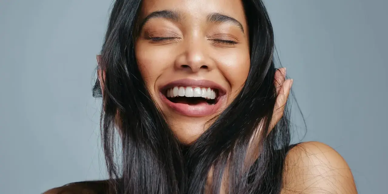 Une femme rit aux éclats, ses yeux fermés. Ses cheveux noirs encadrent son visage radieux, témoignant de la beauté que la vitamine B7 peut apporter.