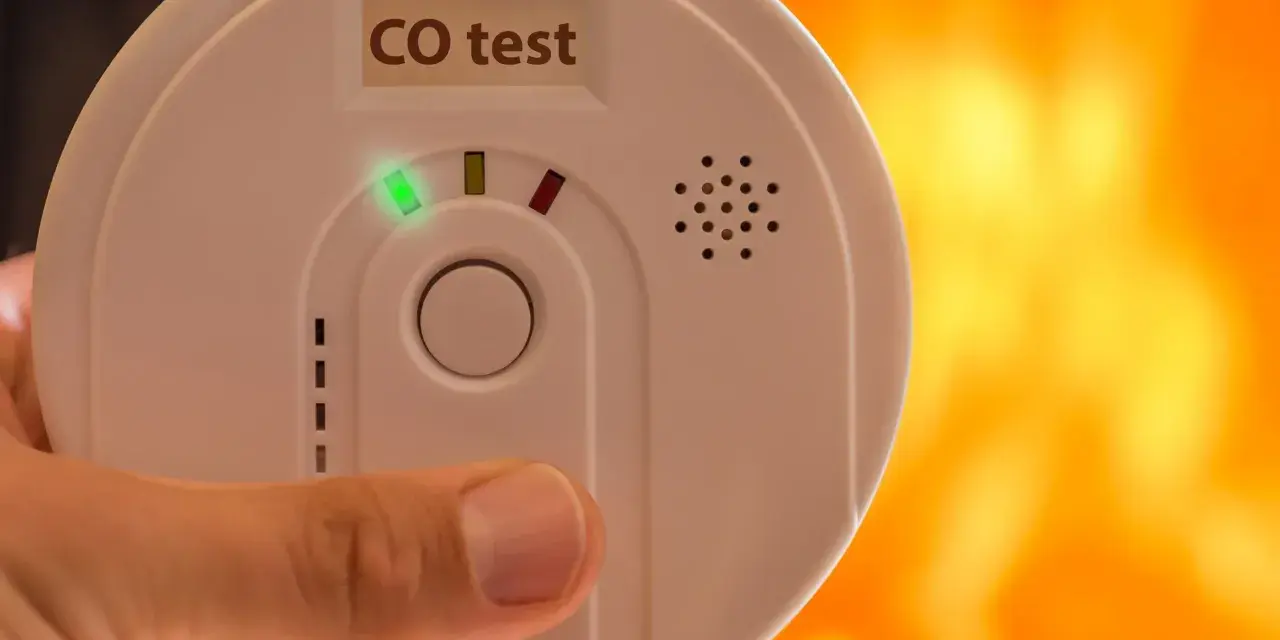 Detektor czadu z zieloną diodą i napisem "CO test" na tle płomieni.