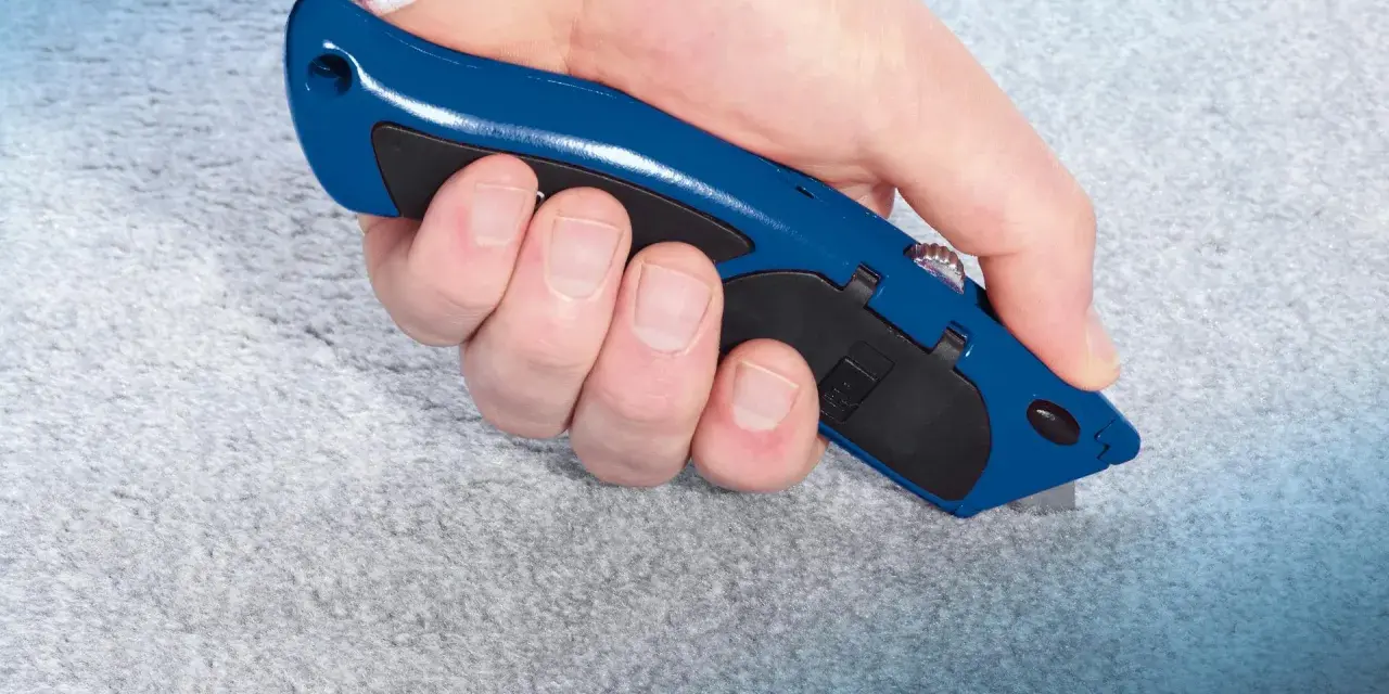 Hand hält blauen Teppichmesser, um grauen Teppich zu schneiden. Präzises Arbeiten beim Teppich verlegen.