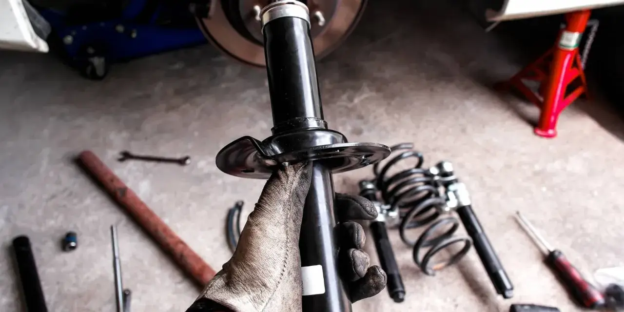 Mano guantata tiene un ammortizzatore nuovo, pronto per sostituire quelli scarichi dell'auto. Attrezzi e molle a vista.