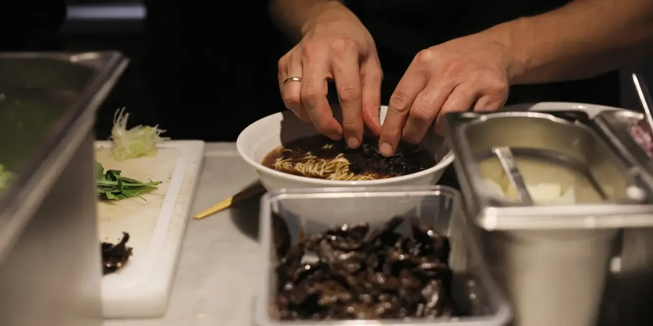 Hände garnieren eine Schüssel mit Ramen-Nudeln in Berlin. Daneben liegen Zutaten bereit.