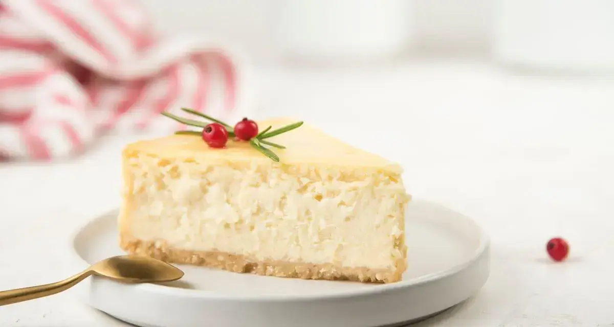 Una fetta di cheesecake con yogurt greco, guarnita con rosmarino e bacche rosse, su un piatto bianco con cucchiaino dorato.