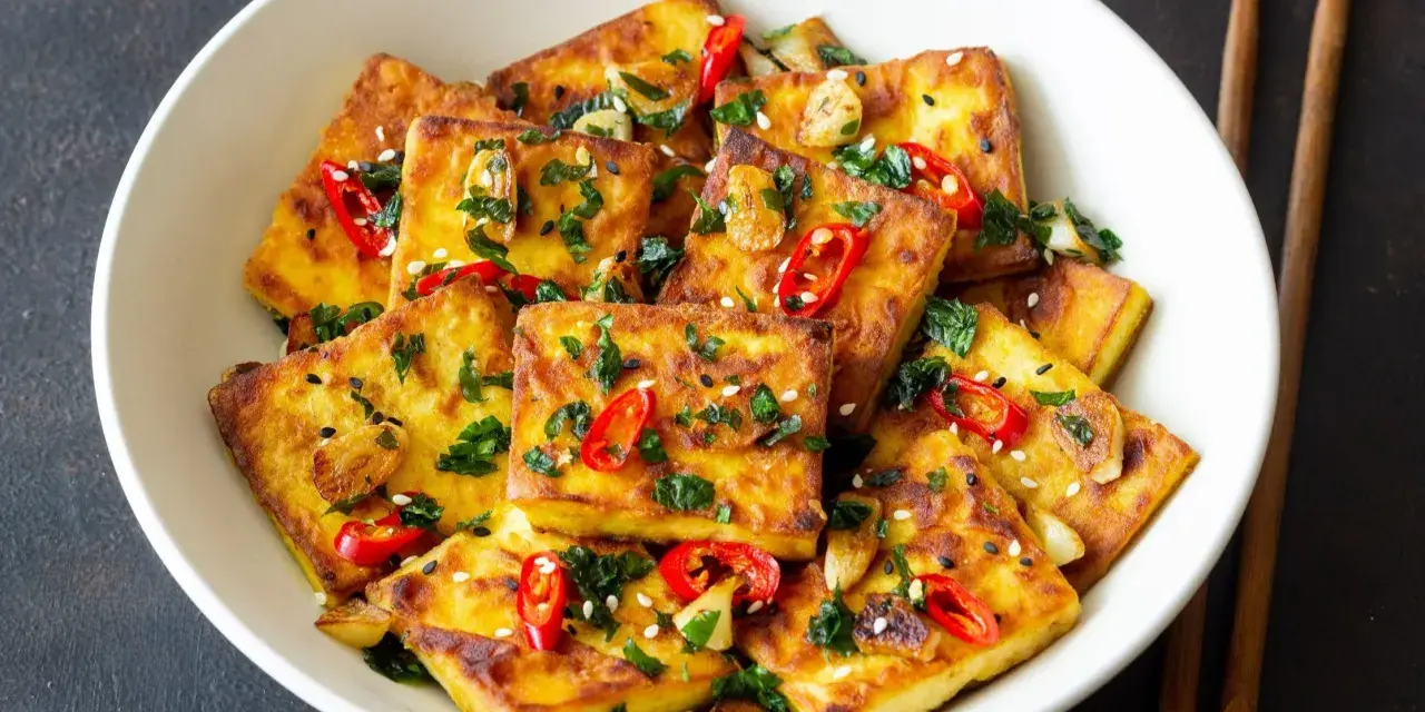 Chrupiące kawałki tofu satay, posypane chili, sezamem i ziołami, podane w białej misce z pałeczkami.