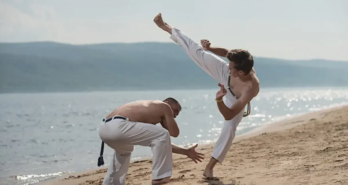 Dwóch mężczyzn ćwiczy capoeirę na plaży. Jeden wykonuje wysokie kopnięcie, drugi jest w pozycji obronnej. Widać ich płynny ginga styl gry.