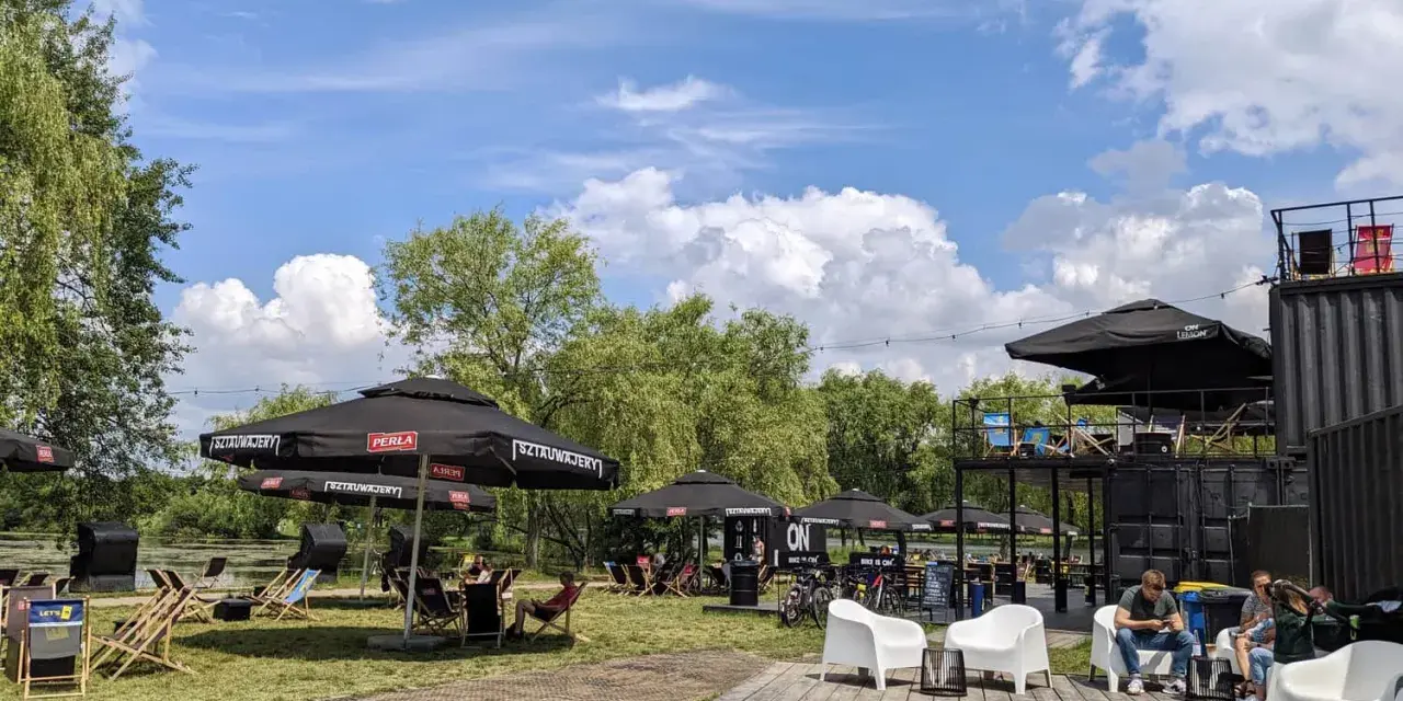 Relaks nad wodą w **Dolinie Trzech Stawów**. Ludzie odpoczywają pod parasolami, a w tle widać zielone drzewa i błękitne niebo z chmurami.