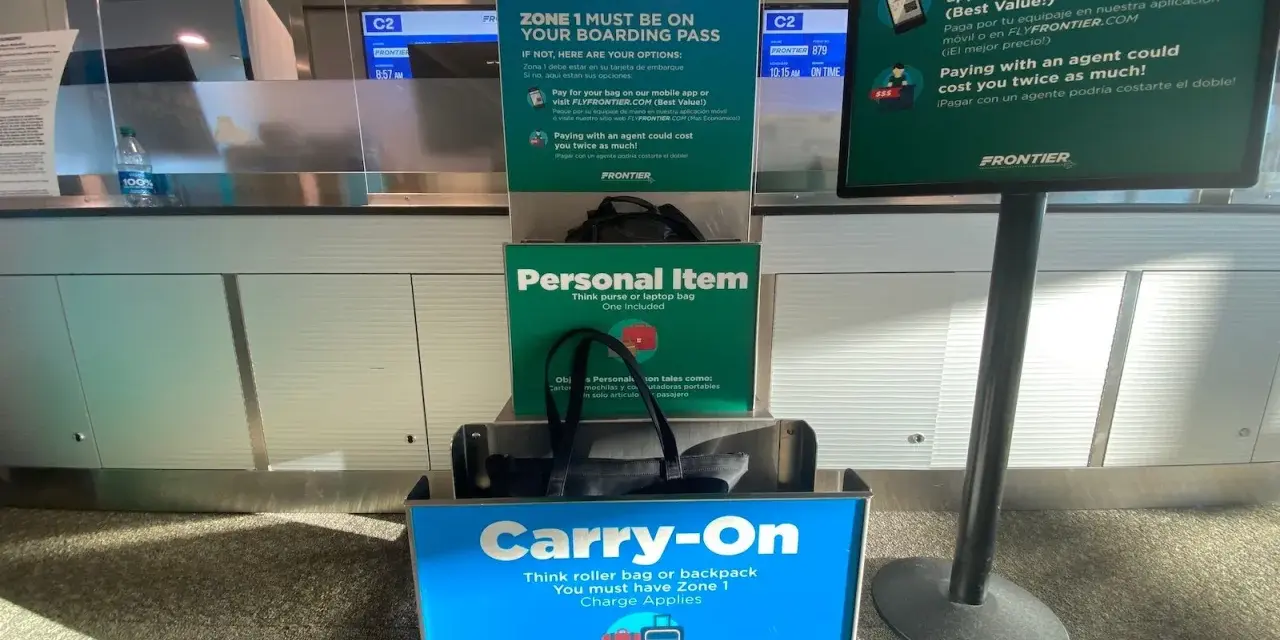 Frontier Airlines personal item size guide at the airport, showing a tote bag fitting in the "Personal Item" box and a roller bag in the "Carry-On" box.