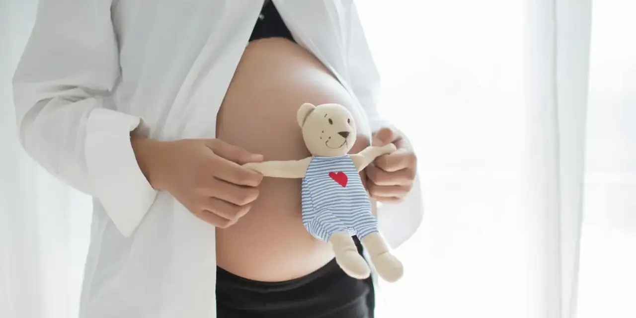 Brzuszek w 21 tygodniu ciąży, trzymający pluszowego misia w pasiastym ubranku z serduszkiem.