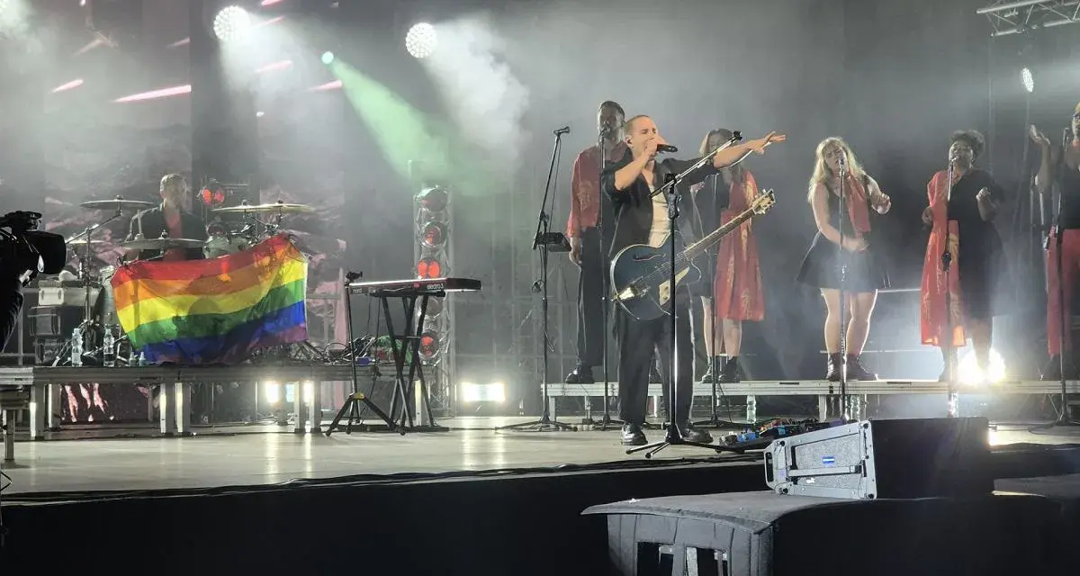 Wokalista z gitarą na scenie, obok chórki i perkusista z tęczową flagą. Koncert podczas festiwalu trzech kultur.