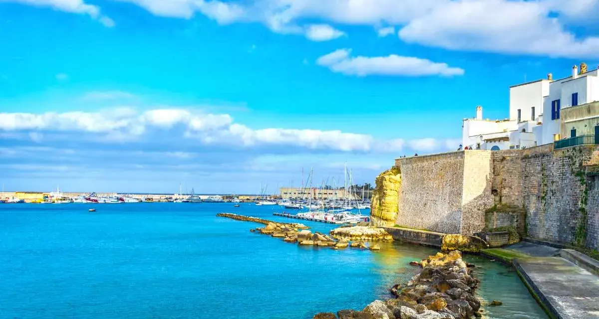 Un incantevole cammino del Salento: un porto con barche a vela, case bianche su scogliere dorate e un mare azzurro sotto un cielo limpido.