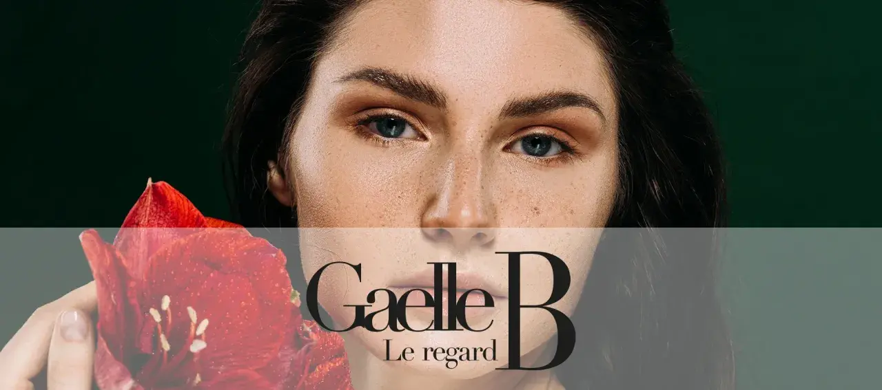 Un visage de femme avec des taches de rousseur naturelles, tenant une fleur rouge. Le regard est intense.