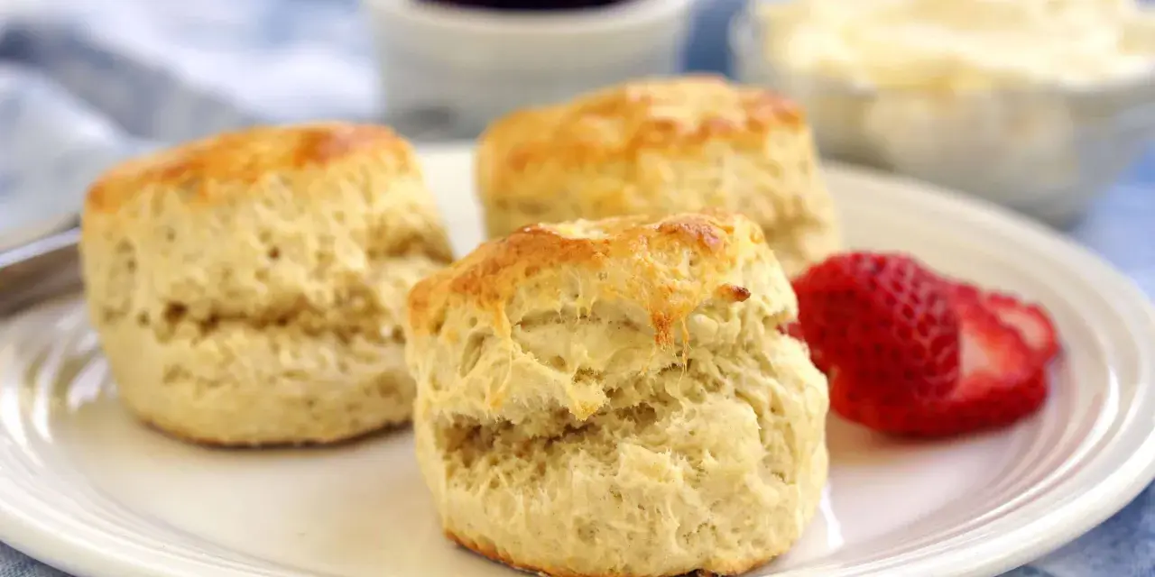 Tre gyllene engelska scones på en vit tallrik med en jordgubbe, sylt och grädde i bakgrunden.