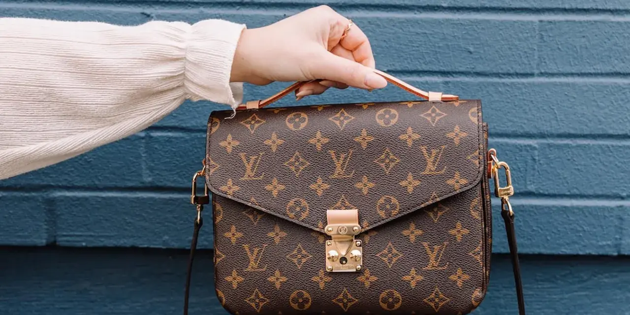 Una mano tiene una borsa Louis Vuitton con il classico monogramma. Un'icona di stile, che ci ricorda dove louis vuitton nasce: dalla maestria artigianale francese.