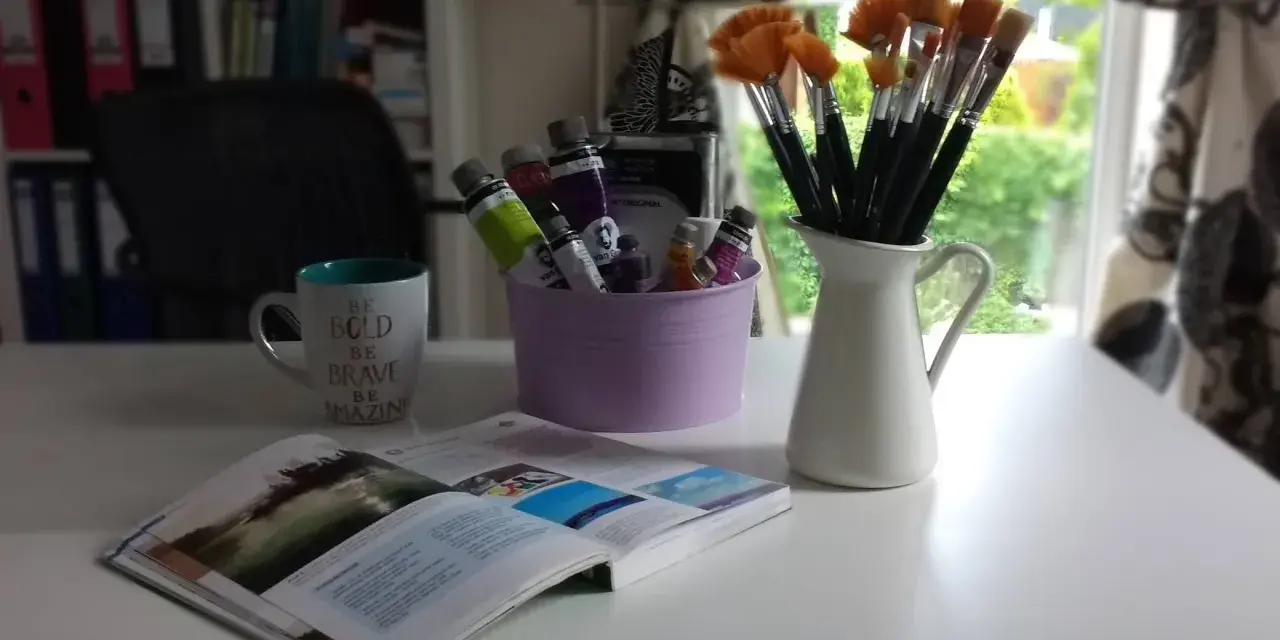 Pędzelki malarskie w dzbanku, tubki farb w fioletowym wiaderku, kubek z napisem i otwarta książka.