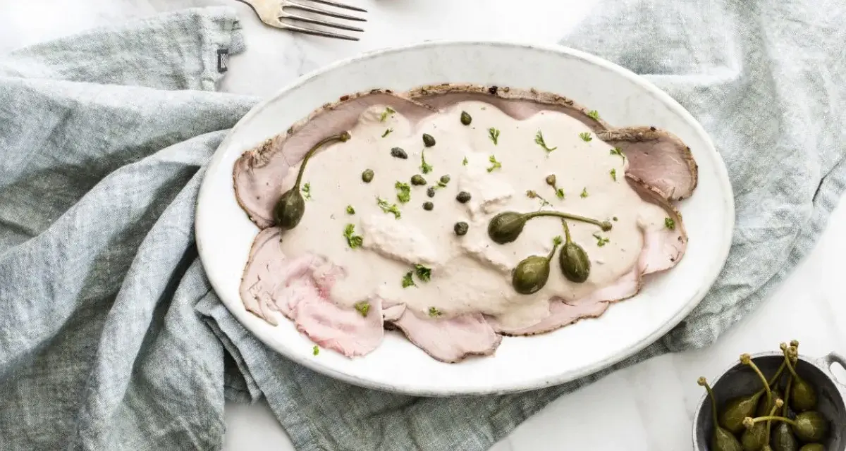 Eleganckie vitello tonnato z kremowym sosem, kaparami i natką pietruszki na białym talerzu, obok srebrnych sztućców i lnianej serwetki.