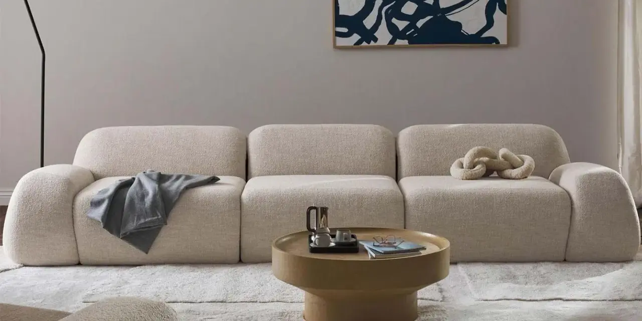 Divano modulare in tessuto bouclé color crema, con cuscino decorativo a forma di catena. Il materiale del divano è morbido e invitante, perfetto per un salotto moderno.