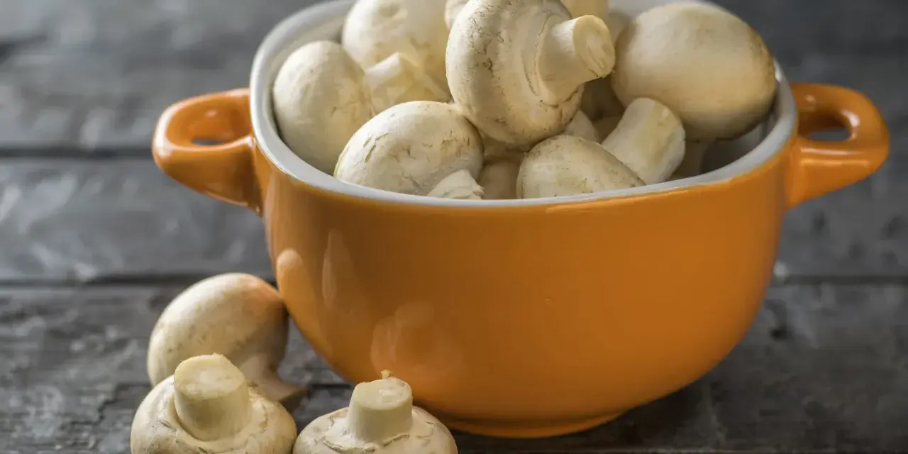 Pomarańczowa miseczka pełna świeżych pieczarek, jakby w ciąży z nowymi smakami. Kilka pieczarek leży obok na drewnianym blacie.