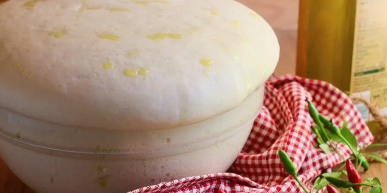 Impasto lievitato per pizza con lievito di birra, soffice e profumato, pronto per essere infornato. Olio e peperoncini completano la scena.