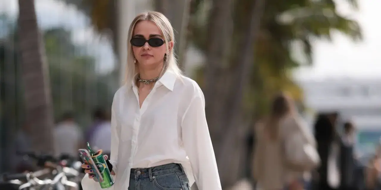 Donna con occhiali da sole, collana, camicia bianca e jeans, tiene in mano una lattina verde e un telefono.