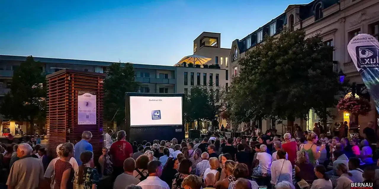 Ein lauer Sommerabend in Berlin: Menschen genießen ein Open-Air-Kino-Erlebnis vor einer Leinwand mit dem Logo "Lux Augenoptik".