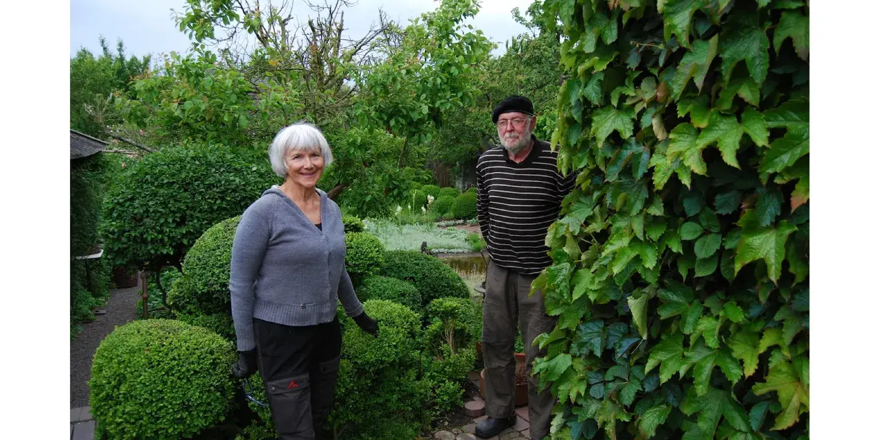 A smiling woman and a man stand in a lush garden with manicured bushes and trees.