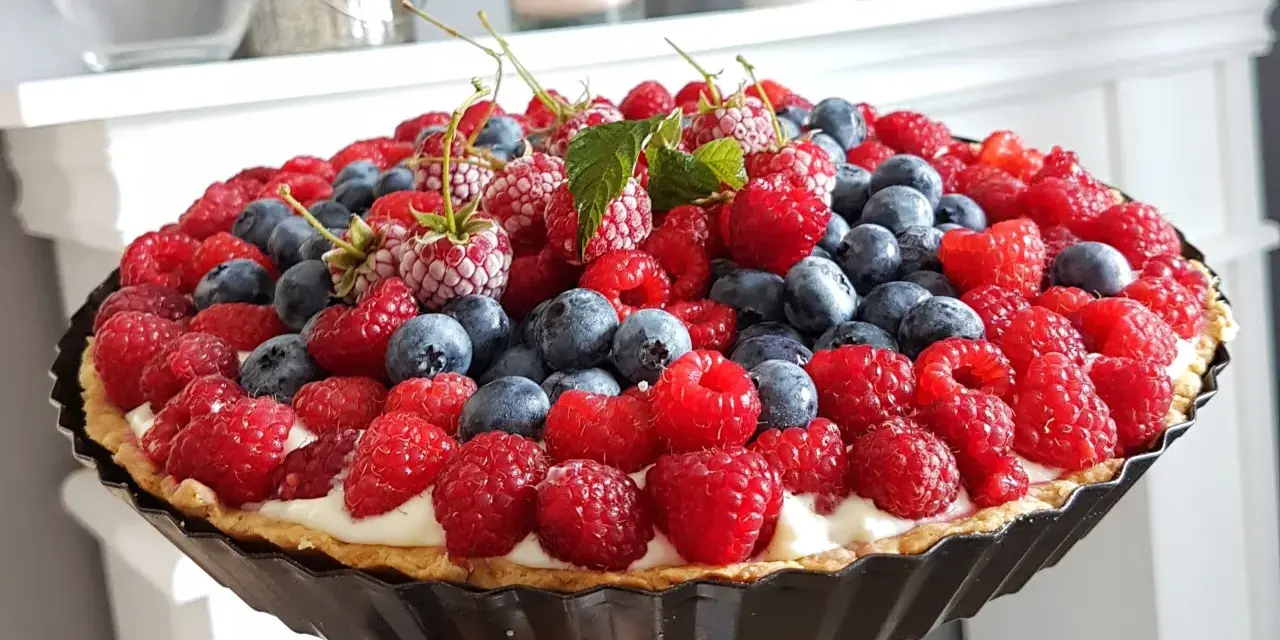 Prosta tarta z budyniem, obficie udekorowana malinami i borówkami, wygląda apetycznie.
