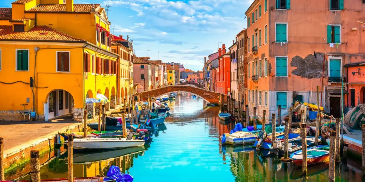 Colorate case affacciate su un canale con barche ormeggiate. Un ponte ad arco collega le due sponde. Un'immagine che ispira su cosa fare a Chioggia.