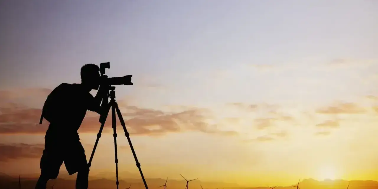 Sylwetka fotografa z aparatem na statywie na tle zachodzącego słońca i farmy wiatrowej. Zastanawiam się, ile bierze fotograf za 18 takich ujęć.
