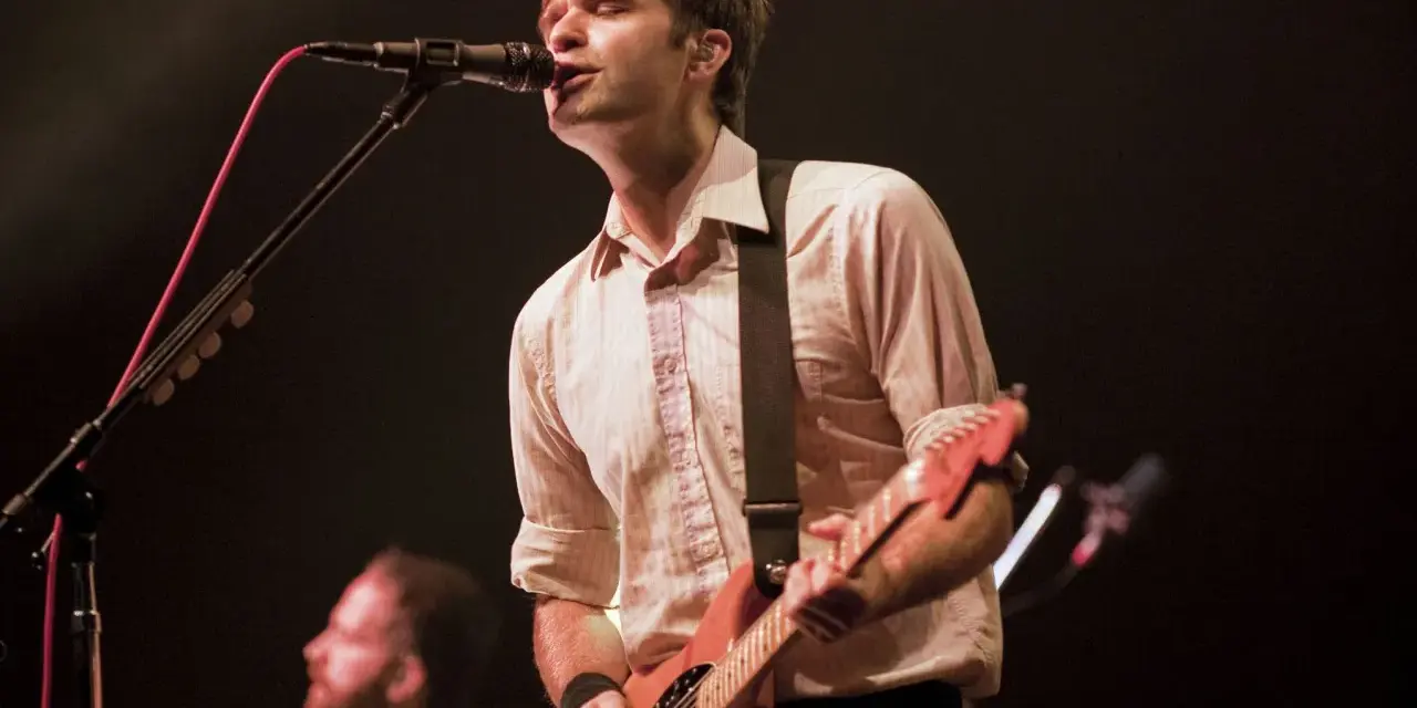 Ben Gibbard chante et joue de la guitare sur scène, éclairé par les projecteurs.