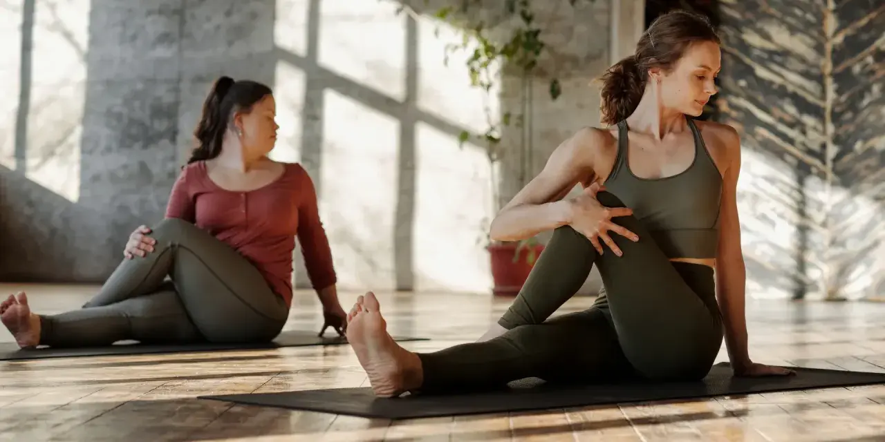Dwie kobiety wykonują skręty tułowia na macie. Delikatne stretching poprawia samopoczucie.