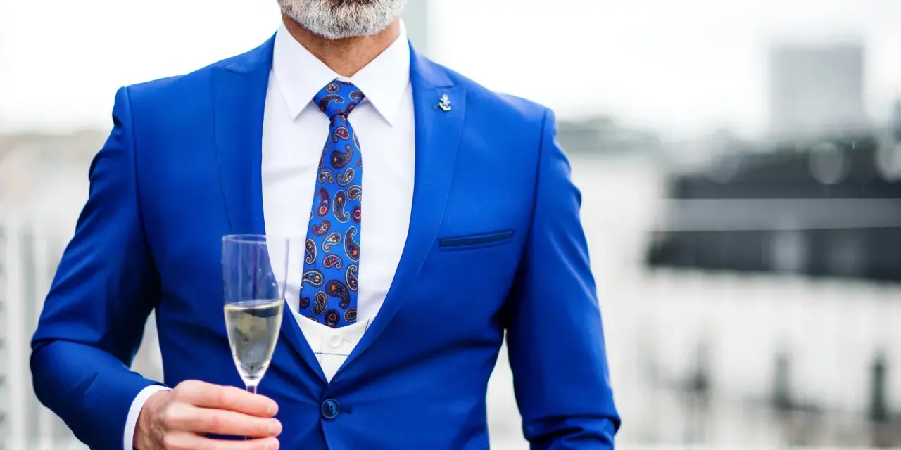 Elegancki mężczyzna w niebieskim garniturze i wzorzystym krawacie, trzymający kieliszek szampana. Idealny strój koktajlowy na specjalne okazje.