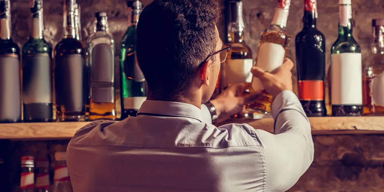 Mann wählt eine Flasche aus, in der reift Whisky in der Flasche nach. Viele Flaschen stehen im Regal.