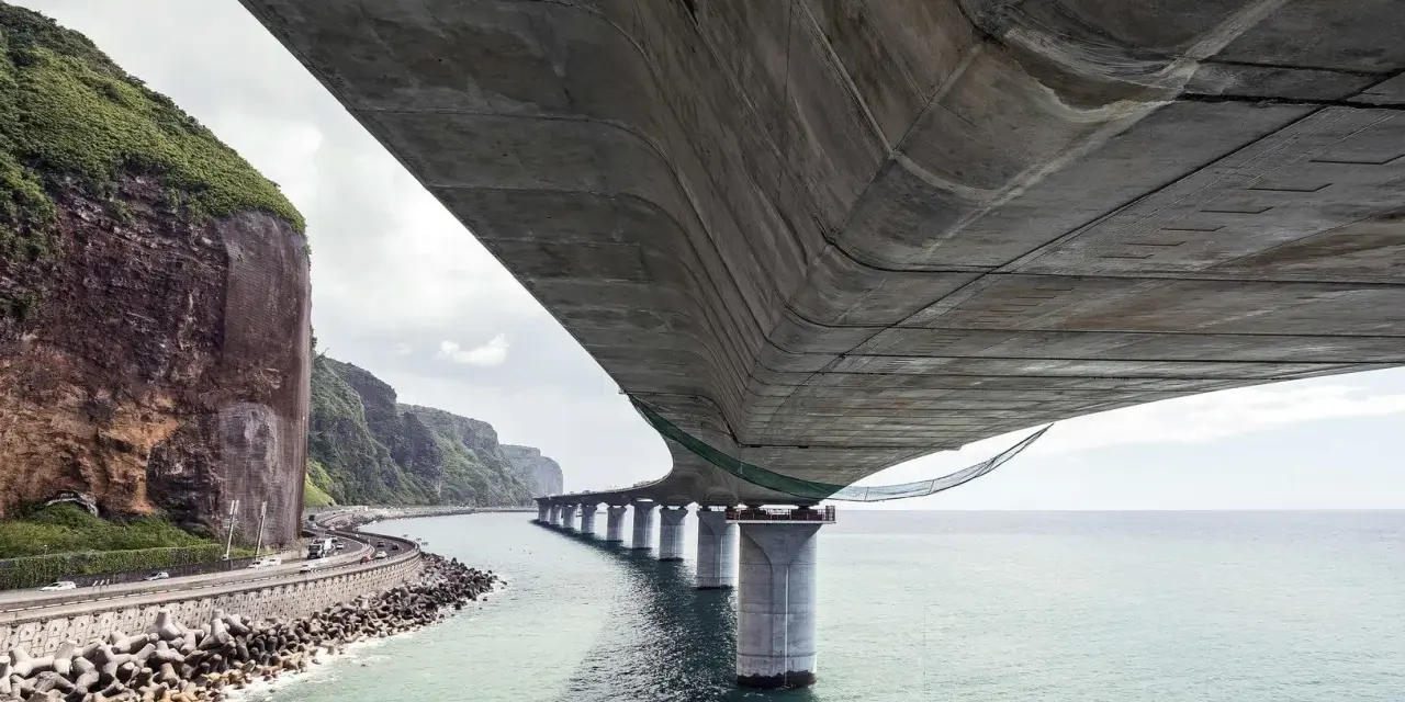 Vue sous un pont en béton, avec la mer, des falaises verdoyantes et une route côtière.
