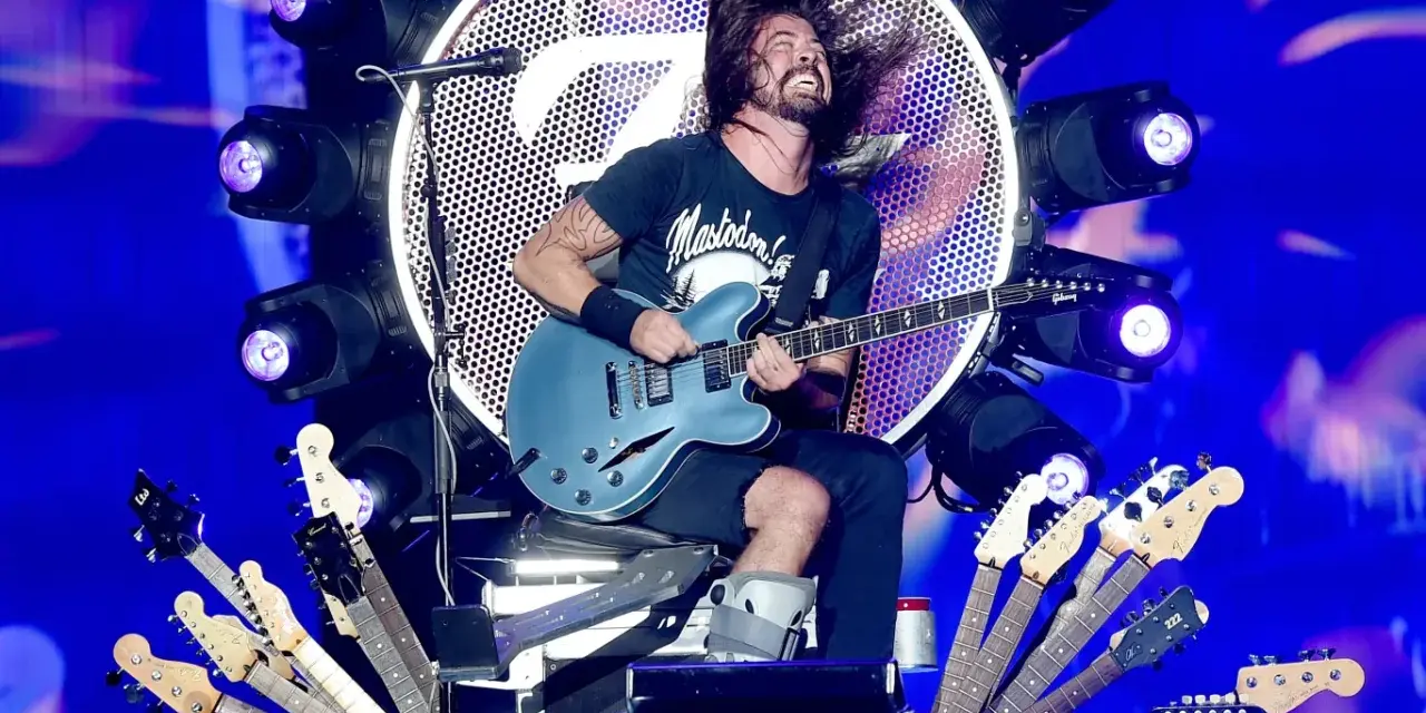 Dave Grohl z zespołem na scenie, szaleje z gitarą. Wokół niego mnóstwo gitar, a w tle wielkie logo i światła.