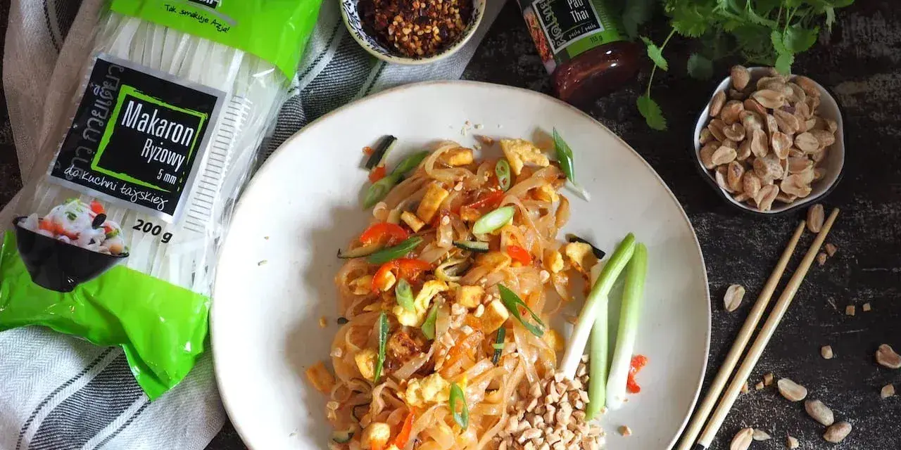 Gotowy Pad Thai przepis na talerzu, obok makaron ryżowy, orzeszki ziemne i sos.