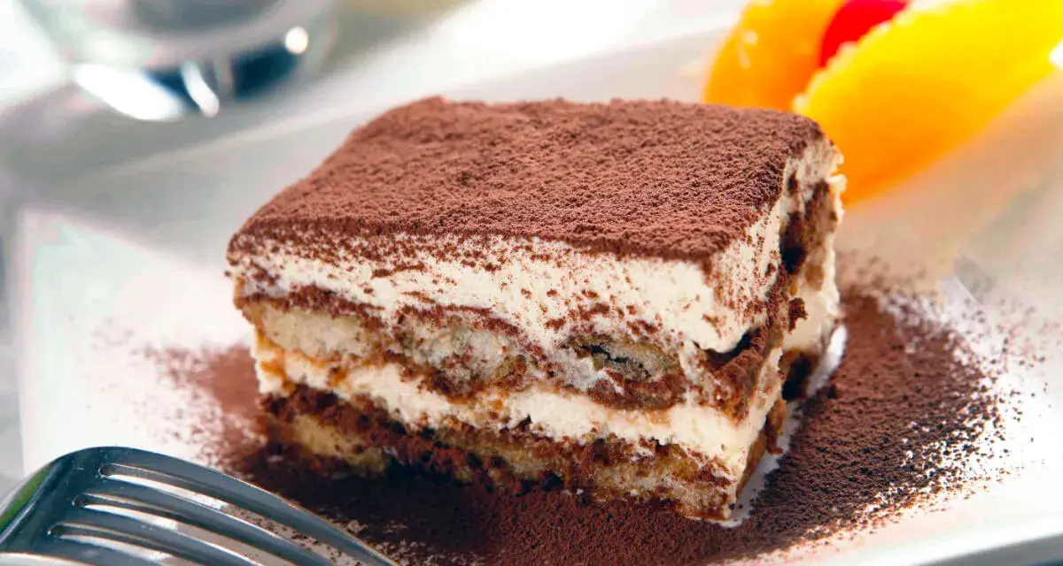 En bit krämig tiramisu, pudrad med kakao, serveras med frukt. Perfekt dryck till tiramisu.