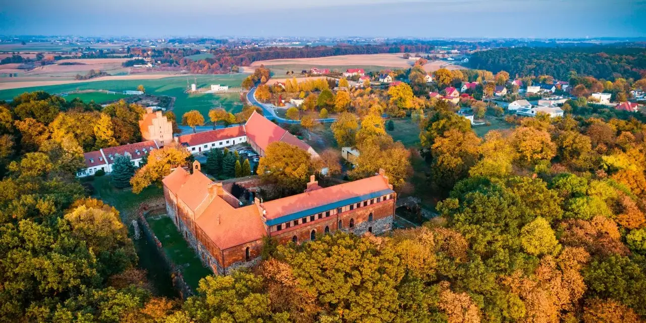 Zamek Bierzgłowski otoczony jesiennym lasem, z czerwonymi dachami i ceglanymi murami, w blasku zachodzącego słońca.