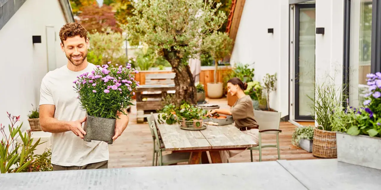 Mann hält Topf mit lila Blumen, um seine Dachterrasse mit Pflanzen zu gestalten. Eine Frau sitzt am Tisch.