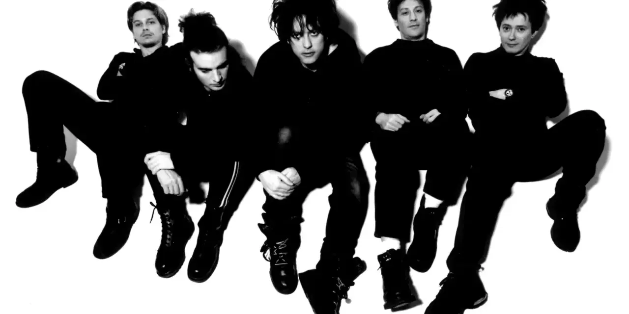 Les membres de The Cure, cinq silhouettes sombres aux coiffures distinctives, posent en groupe, l'air pensif.