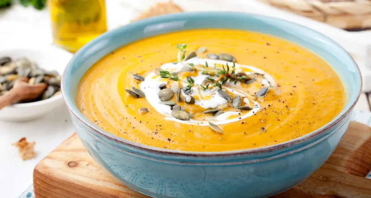 Vellutata di zucca cremosa, guarnita con panna, semi di zucca e timo, servita in una ciotola azzurra su un tagliere di legno.