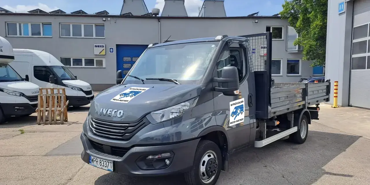Szary Iveco z otwartą skrzynią ładunkową, idealny do wynajem wywrotki do 3,5 tony. Na drzwiach logo "Wypożyczalnia Moto-Partner".