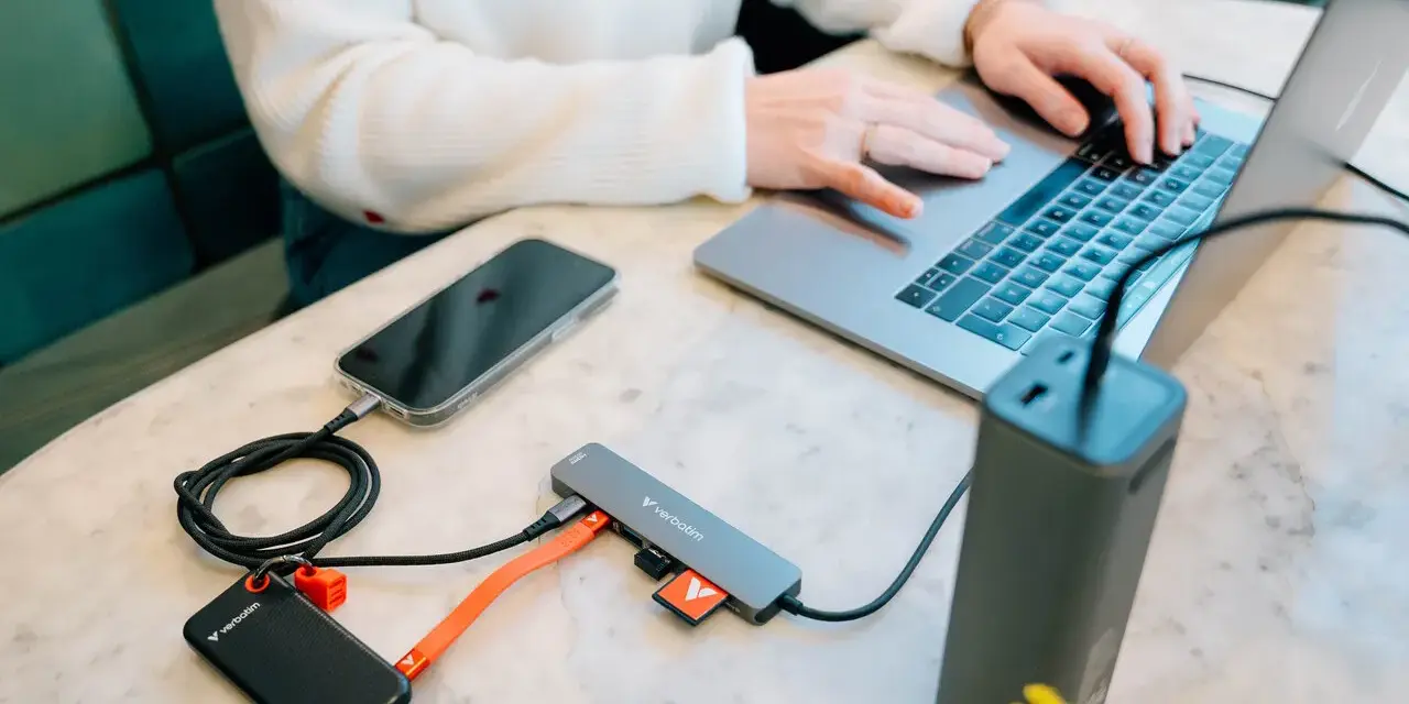 En person använder en laptop med en hubb ansluten till en telefon, extern hårddisk och powerbank. Hubben har usb 3.2-portar.