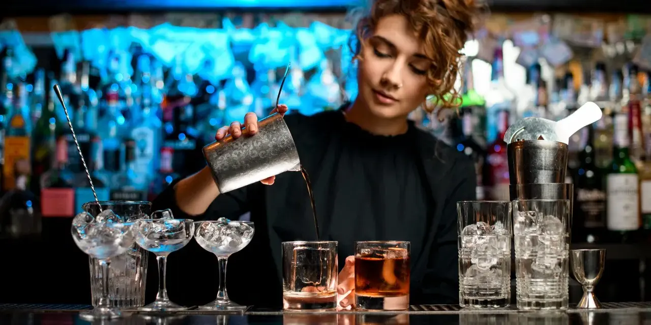 Barman nalewa drink do szklanki na tle butelek i niebieskiego światła.
