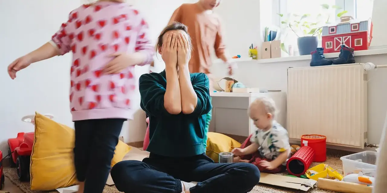 Chaos im Kinderzimmer. Eine Mutter versteckt ihr Gesicht in den Händen, während ihre Kinder spielen. Trotz des Trubels ist die liebe zu kindern spürbar.