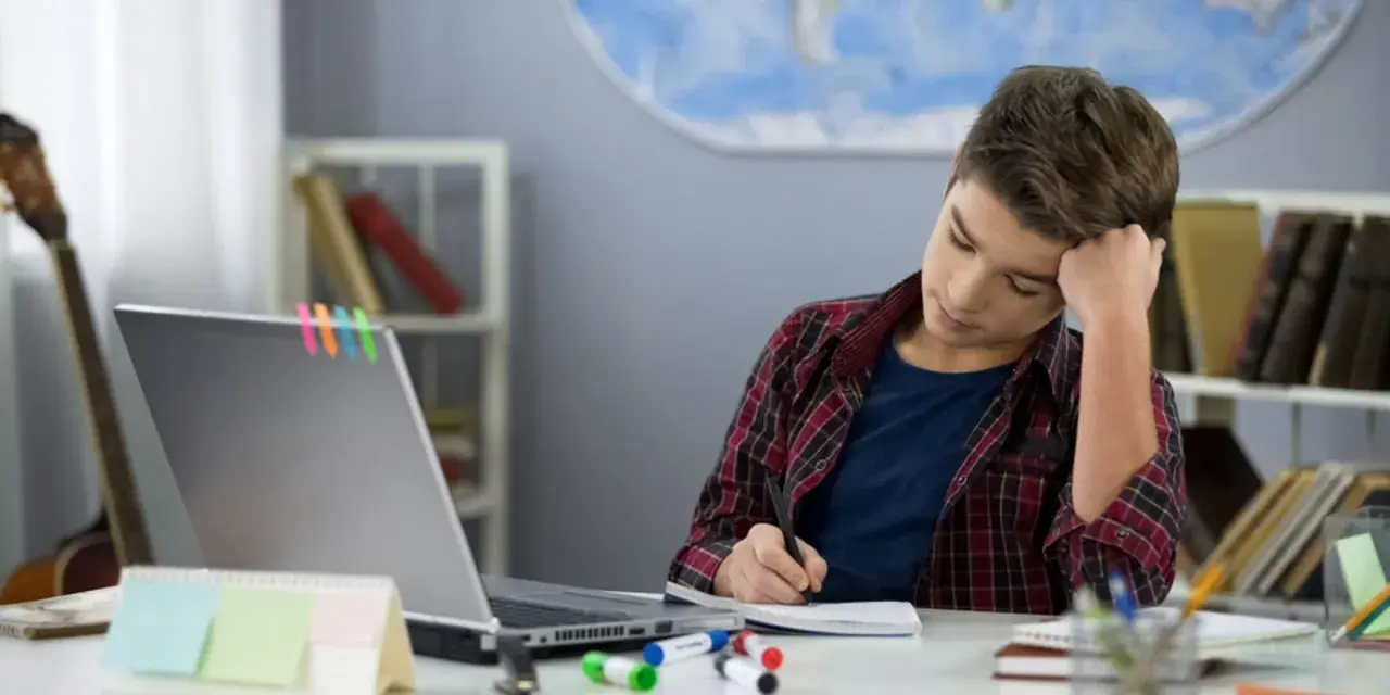 Ein Junge sitzt frustriert am Schreibtisch und macht seine hausaufgaben. Ein Laptop und bunte Stifte liegen bereit.