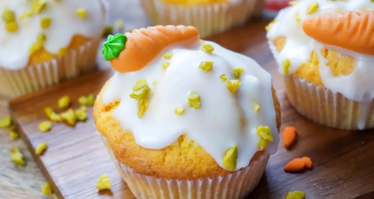 Muffins mit weißer Glasur, gehackten Pistazien und Marzipan-Karotten auf Holzbrett.