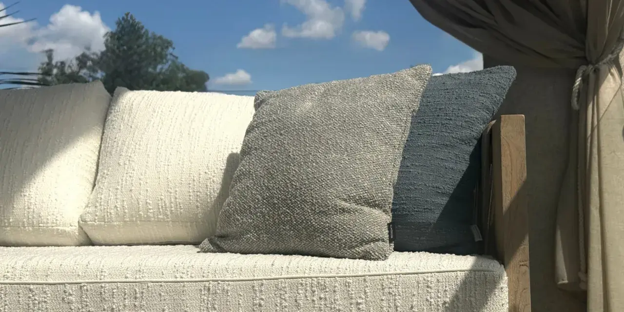 Wygodna sofa z poduszkami, osłonięta zasłonami, stoi na tle błękitnego nieba. Jej rama wykonana jest z pięknego, naturalnego **drewna tekowego**.